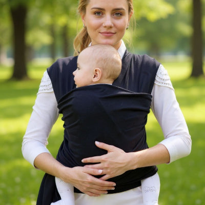Maman souriante tenant bébé en écharpe de portage dans un parc, moment de proximité et confort physiologique.