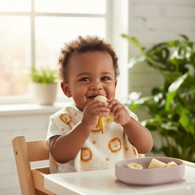 Bébé africain en chaise haute mangeant une banane, portant un bavoir bébé manche longue en coton motif lion, bavoir coton bébé absorbant pour diversification alimentaire.
