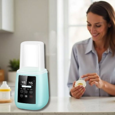 Chauffe biberon multifonction 6-en-1 couleur turquoise posé sur un plan de travail de cuisine, à côté d'une maman qui nettoie une tétine. Chauffage précis à 45°C affiché à l'écran digital.
