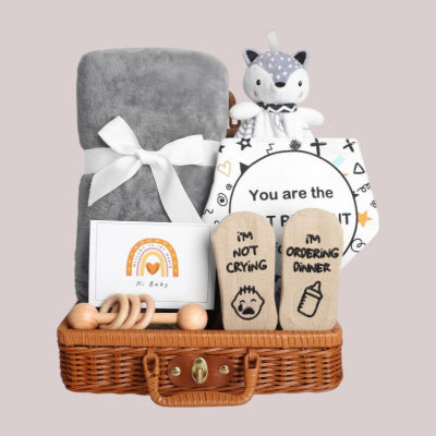 Coffret Naissance Bébé avec couverture grise, doudou renard, bavoir illustré, chaussettes humoristiques, carte de bienvenue et hochet en bois naturel, présenté dans un panier en osier.