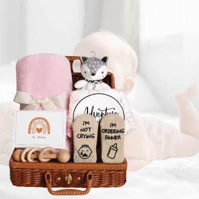 Coffret naissance bébé placé sur un lit aux côtés d’un bébé fille, avec couverture rose, chaussettes humoristiques et doudou en peluche dans un panier en osier.