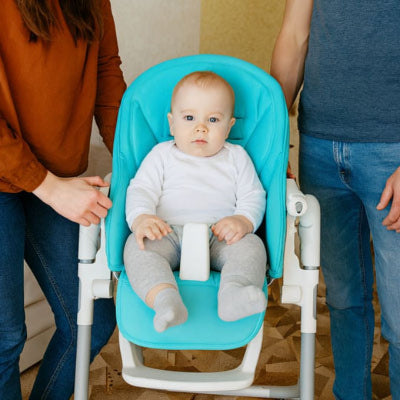 Bébé assis sur coussin chaise haute bébé turquoise avec housse protectrice et harnais 5 points, parents veillant à la sécurité pendant le repas.