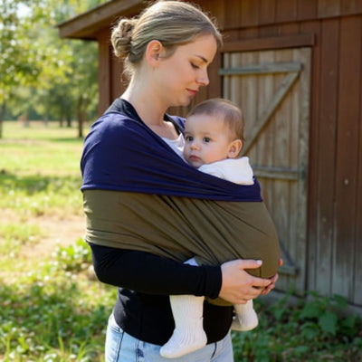 Maman portant son bébé dans une écharpe de portage kaki et bleu marine à l’extérieur, devant un cabanon en bois, dans un jardin verdoyant et paisible.