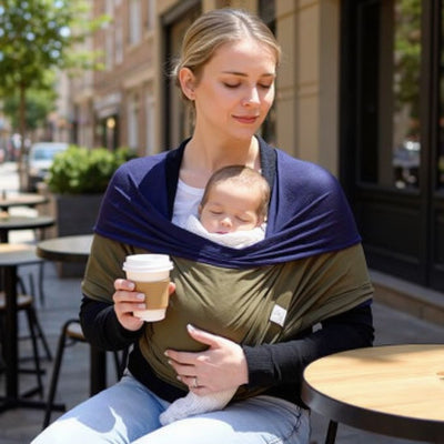 Maman assise en terrasse d’un café tenant son bébé endormi dans une écharpe de portage kaki et bleu marine, boisson chaude à la main, ambiance urbaine tranquille.