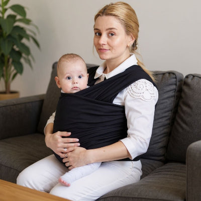 Jeune maman assise sur un canapé avec bébé porté en écharpe de portage noire, position sécurisée idéale pour allaiter à la maison.