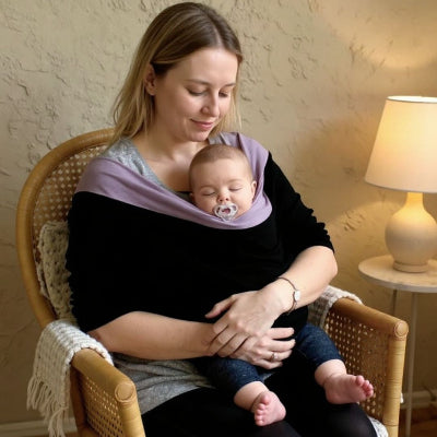 Maman installée dans un fauteuil en rotin tenant son bébé endormi dans une écharpe de portage noir et lavande, lampe d’ambiance et plaid en fond, intérieur cosy.
