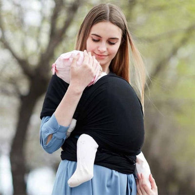 Maman tenant tendrement son bébé contre elle dans une écharpe de portage noire, en extérieur sous des arbres en fleurs, moment de douceur printanière.