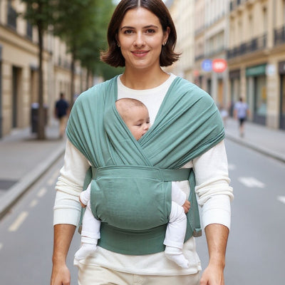 Maman souriante portant son bébé en écharpe de portage vert sauge dans la rue, illustrant le confort et la praticité du portage physiologique au quotidien.