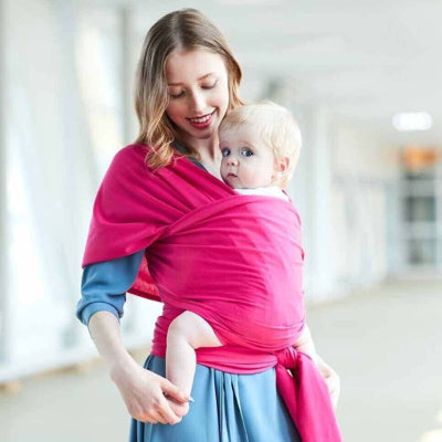 Maman souriante en intérieur portant un bébé blond aux yeux bleus dans une écharpe de portage rose, ambiance lumineuse et vêtement bleu pastel.