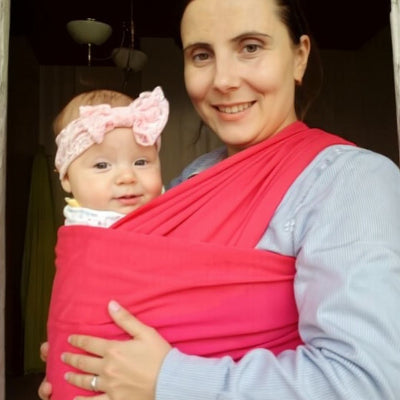 Maman souriante tenant bébé heureux en écharpe de portage rose, en intérieur, illustrant le confort et le lien affectif renforcé.