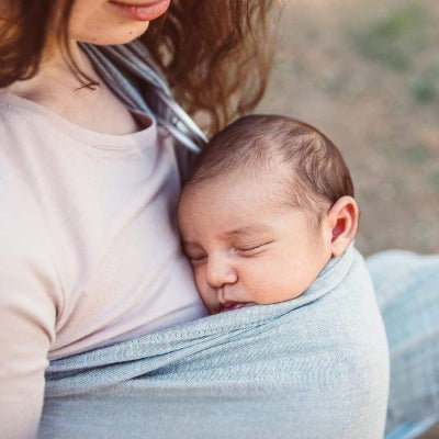 Gros plan sur un nouveau-né qui dort paisiblement contre sa maman dans une écharpe de portage sans nœud, tissu coton respirant.