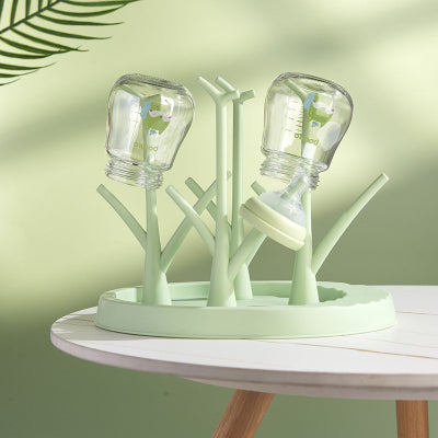 Égouttoir biberon vert clair sur table ronde blanche, avec deux biberons positionnés tête en bas, dans une ambiance zen et naturelle.