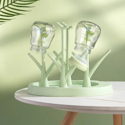 Égouttoir biberon vert clair sur table ronde blanche, avec deux biberons positionnés tête en bas, dans une ambiance zen et naturelle.