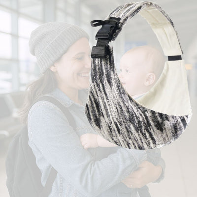 Maman souriante tenant son bébé à bras à l’aéroport, illustrant le besoin d’une écharpe de portage pour voyager plus sereinement.