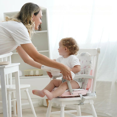 Rehausseur chaise pliable bébé rose en cours d’installation par une maman, siège pratique et sécurisé pour transformer une chaise classique en chaise bébé de table.
