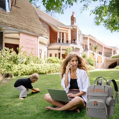 Sac à dos à langer BabyNest™ en gris élégant, idéal pour des journées actives, avec une maman travaillant depuis un jardin tout en surveillant son enfant, reflétant confort et praticité dans chaque détail.