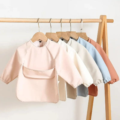 Collection de bavoirs bébé manche longue imperméables suspendus sur cintres, lot bavoir bébé fille et garçon avec manches, disponible en coloris pastels assortis.