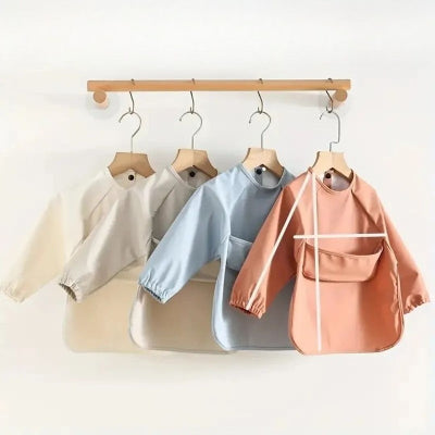 Ensemble de bavoirs bébé manche longue crème, bleu ciel et terracotta accrochés sur cintres, grand bavoir bébé avec manches et récupérateur pour repas propres et faciles.