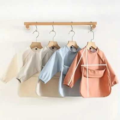 Ensemble de bavoirs bébé manche longue crème, bleu ciel et terracotta accrochés sur cintres, grand bavoir bébé avec manches et récupérateur pour repas propres et faciles.