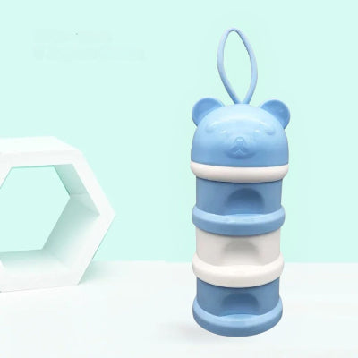 Boite doseuse lait bleue en forme d’ourson avec 3 compartiments empilables et anse souple, posée sur une surface blanche, idéale pour stocker du lait infantile en déplacement.