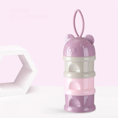 Boite doseuse lait en forme d’ourson violet avec trois compartiments empilables, posée sur une table blanche, fond pastel rose.