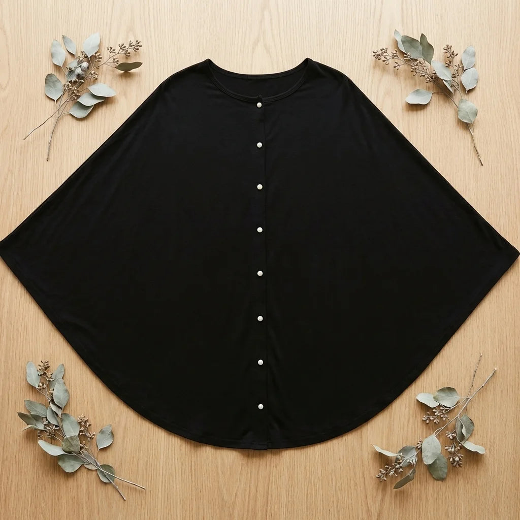 Cape pour allaitement Solène noire étalée à plat sur bois, 7 boutons nacre blancs visibles, coupe semi-circulaire, branches eucalyptus