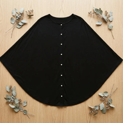 Cape pour allaitement Solène noire étalée à plat sur bois, 7 boutons nacre blancs visibles, coupe semi-circulaire, branches eucalyptus