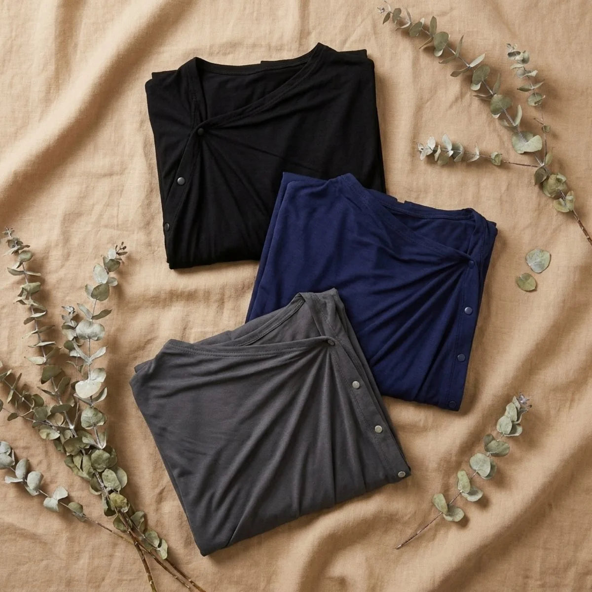 Flatlay cape pour allaitement Solène™ — trois coloris noir, marine et gris sur tissu lin et branches d'eucalyptus