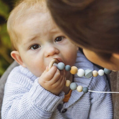 Bébé tenant un collier d’allaitement avec anneau en bois et perles silicone, parfait pour calmer les gencives douloureuses et éveiller les sens.