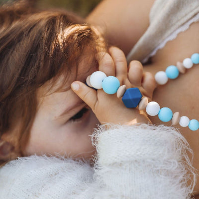 Bébé en portage tenant un collier d’allaitement bleu ciel et blanc, conçu pour occuper les mains curieuses et soulager les gencives.