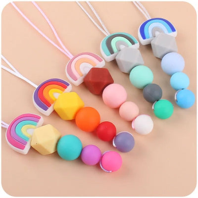 Gamme de colliers d’allaitement avec pendentifs en forme d’arc-en-ciel et perles colorées, conçus pour occuper les mains de bébé et apaiser les douleurs dentaires pendant les moments de proximité.