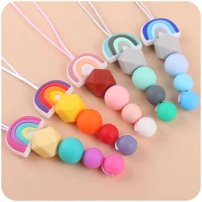 Gamme de colliers d’allaitement avec pendentifs en forme d’arc-en-ciel et perles colorées, conçus pour occuper les mains de bébé et apaiser les douleurs dentaires pendant les moments de proximité.