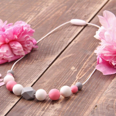 Collier d’allaitement rose et blanc posé entre des fleurs, accessoire sensoriel et esthétique à offrir ou porter pour distraire bébé