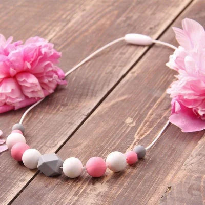 Collier d’allaitement rose et blanc posé entre des fleurs, accessoire sensoriel et esthétique à offrir ou porter pour distraire bébé