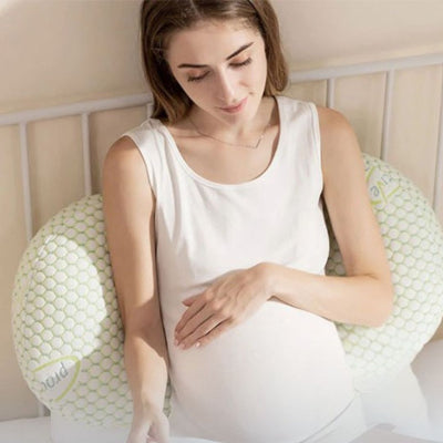 Femme enceinte assise dans son lit, adossée à un coussin de grossesse en forme de U pour un moment de lecture détendu et confortable.