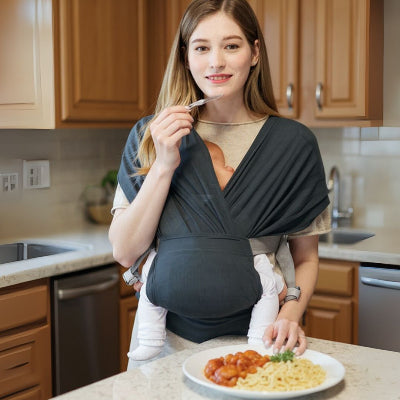 Jeune maman allaitant son bébé dans une écharpe de portage tout en préparant un repas, en cuisine.