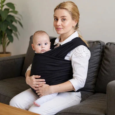 Jeune maman assise sur un canapé avec bébé porté en écharpe de portage noire, position sécurisée idéale pour allaiter à la maison.