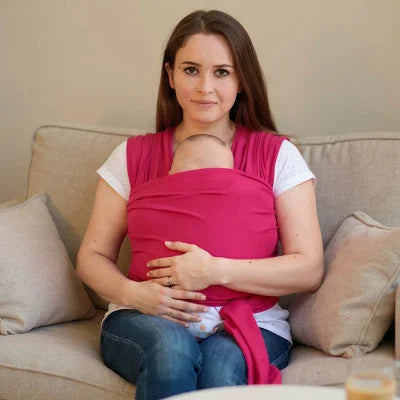 Maman assise sur un canapé beige allaitant son bébé porté contre elle dans une écharpe de portage rose, position physiologique et ambiance cocooning.