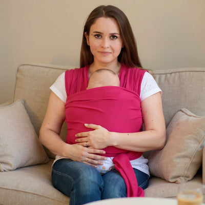 Maman assise sur un canapé beige allaitant son bébé porté contre elle dans une écharpe de portage rose, position physiologique et ambiance cocooning.