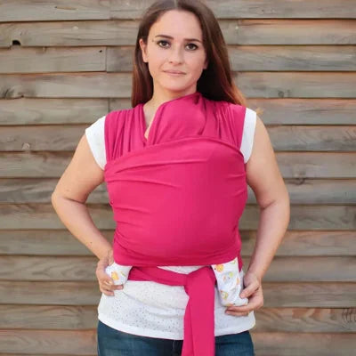 Femme debout face caméra portant son bébé dans une écharpe de portage rose devant un mur en bois, position ventrale serrée et tissu extensible.