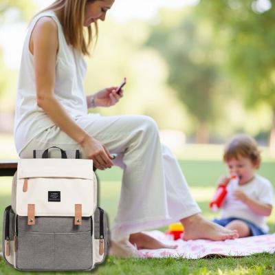 Une maman assise en plein pique-nique avec son enfant dans un parc, accompagnée du Sac à dos à langer ÉlégancePlus™ Écru Élégance posée à proximité. Ce sac allie praticité et style pour des sorties organisées, avec de nombreux compartiments pour les affaires de bébé et un design élégant pour les parents.