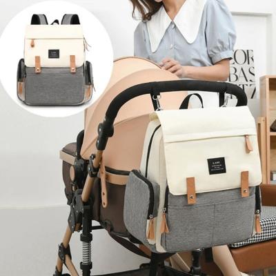 Sac à dos à langer ÉlégancePlus™ Écru Élégance fixé sur une poussette beige avec une maman élégante en arrière-plan. Idéal pour les sorties familiales, il allie design moderne et praticité pour les parents.