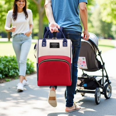 Sac à langer bébé rouge transporté à la main, adapté pour une sortie avec poussette en plein air, illustrant sa polyvalence et son design élégant.