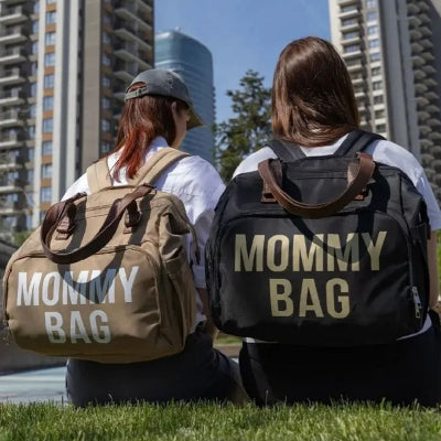 Deux mamans assises sur l'herbe dans un parc urbain, portant des sacs à langer Beige Chic et Noir Sophistiqué, parfaits pour une sortie organisée avec bébé.