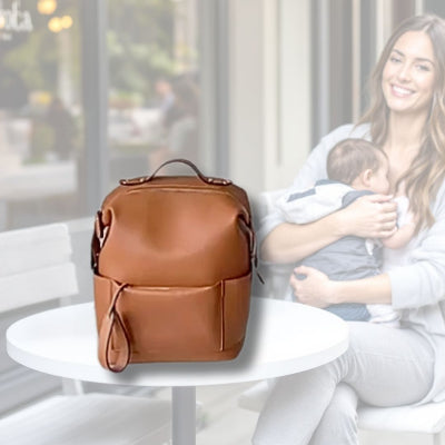 Sac à langer couleur caramel chic sur une table dans un intérieur lumineux et confortable, avec une maman allaitant son bébé en arrière-plan, soulignant ses finitions en cuir vegan et ses multiples compartiments.