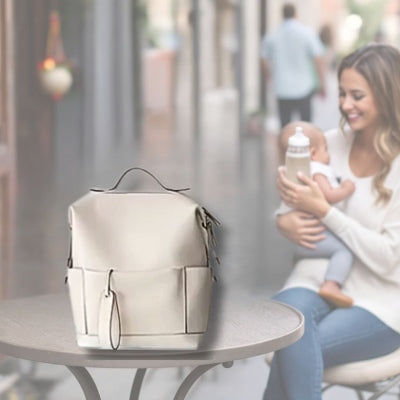 Sac à langer couleur ivoire pureté placé sur une table lors d'une balade en ville, avec une maman donnant un biberon à son bébé en arrière-plan, parfait pour une utilisation quotidienne grâce à son design fonctionnel et chic.