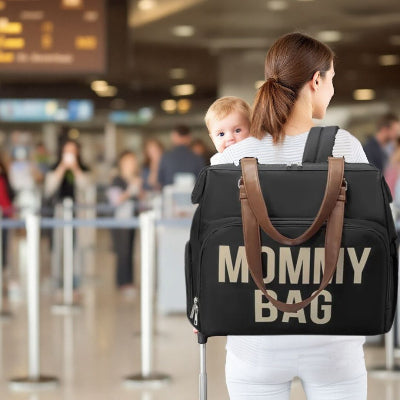 Une maman avec un sac à langer noir sophistiqué porté sur ses épaules dans un aéroport, idéal pour organiser les affaires de bébé en voyage.