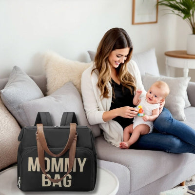 Une maman souriante joue avec son bébé dans un intérieur chaleureux, le sac à langer Noir Sophistiqué posé sur une table, prêt pour la prochaine sortie.