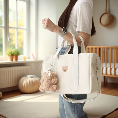 Sac à langer blanc crème avec un motif ourson tendre, présenté dans une chambre lumineuse avec un berceau en arrière-plan, idéal pour les mamans modernes.