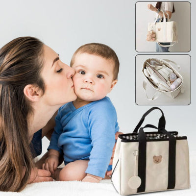 Une maman embrassant tendrement son bébé, avec un sac à langer ourson tendre visible au premier plan. Le sac offre un design élégant en blanc cassé avec des poignées noires, parfait pour transporter les essentiels de bébé.